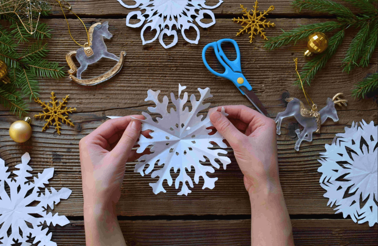 Hands folding a paper snowflake among Christmas ornaments and craft supplies
