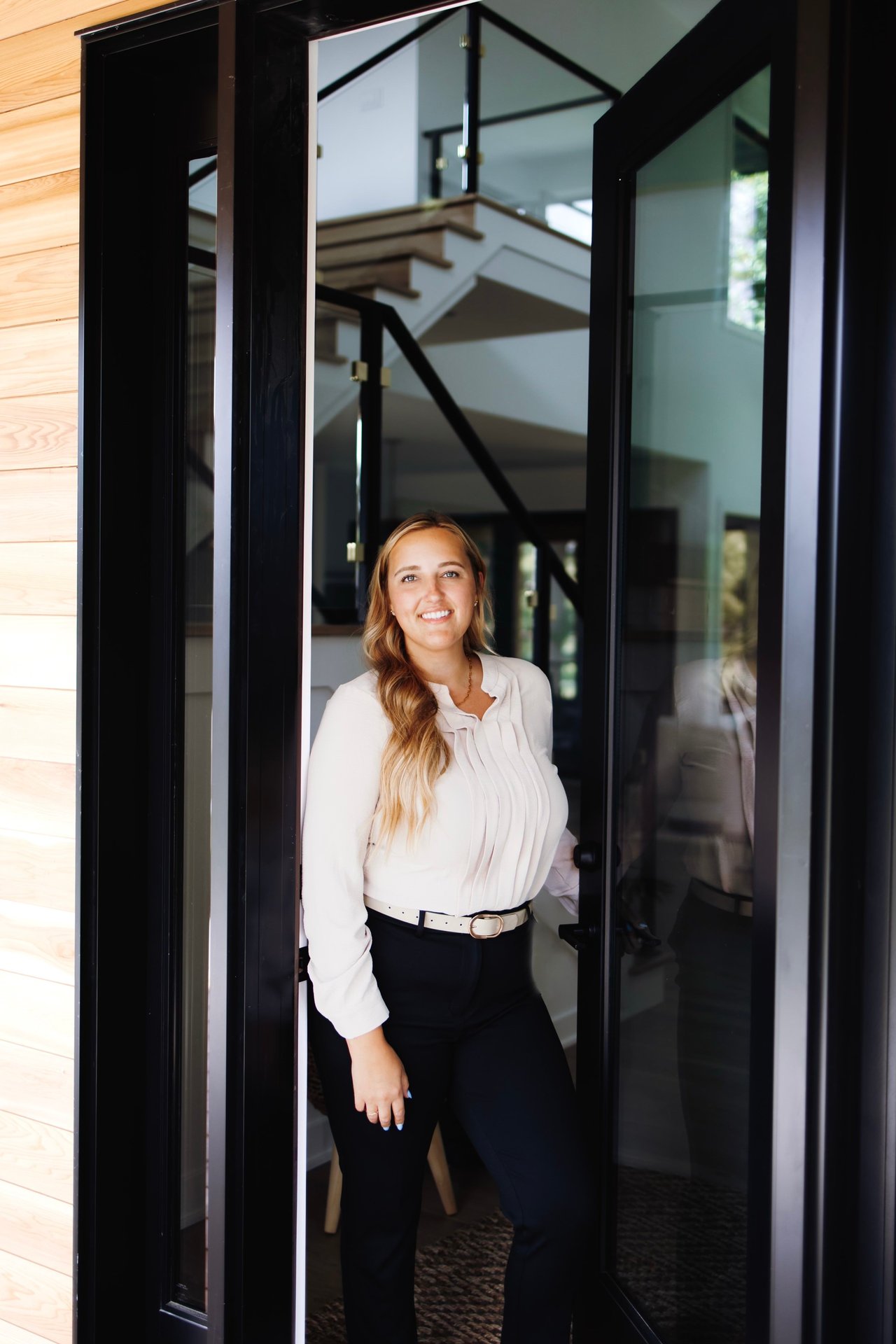 Woman in a white blouse standing in an open glass doorway with a staircase behind her.
