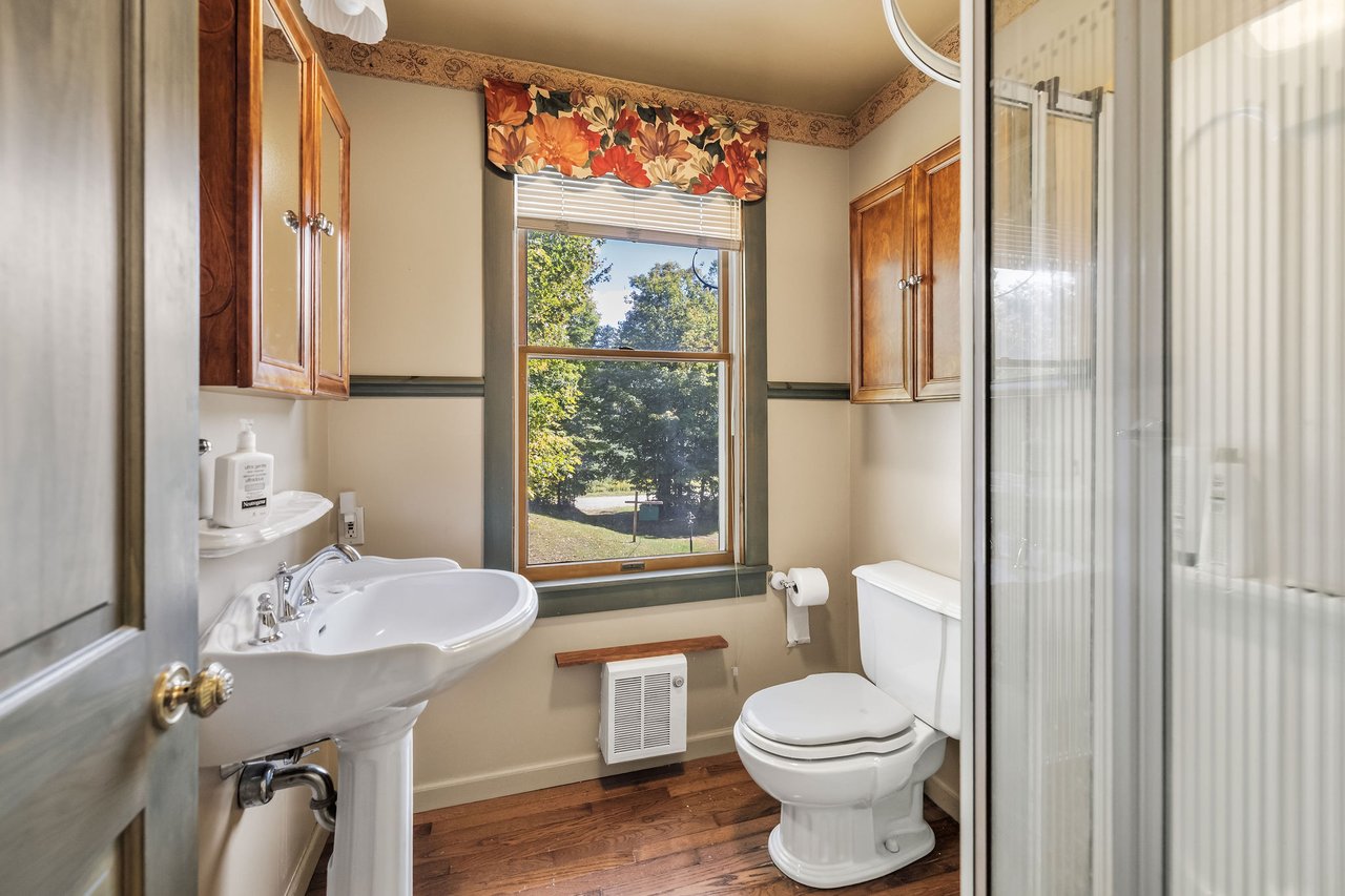 Bathroom with a pedestal sink, glass shower enclosure, and window overlooking leafy grounds.