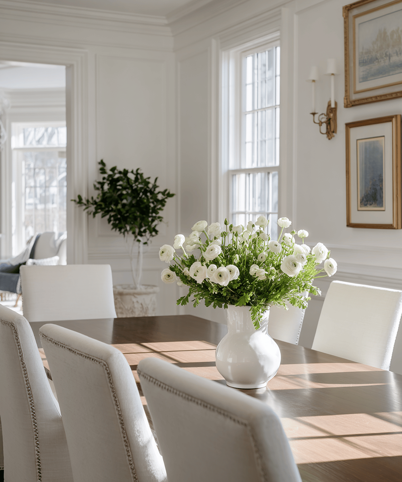 Bright dining room with white chairs, tall windows, and a vase of white flowers on the table.