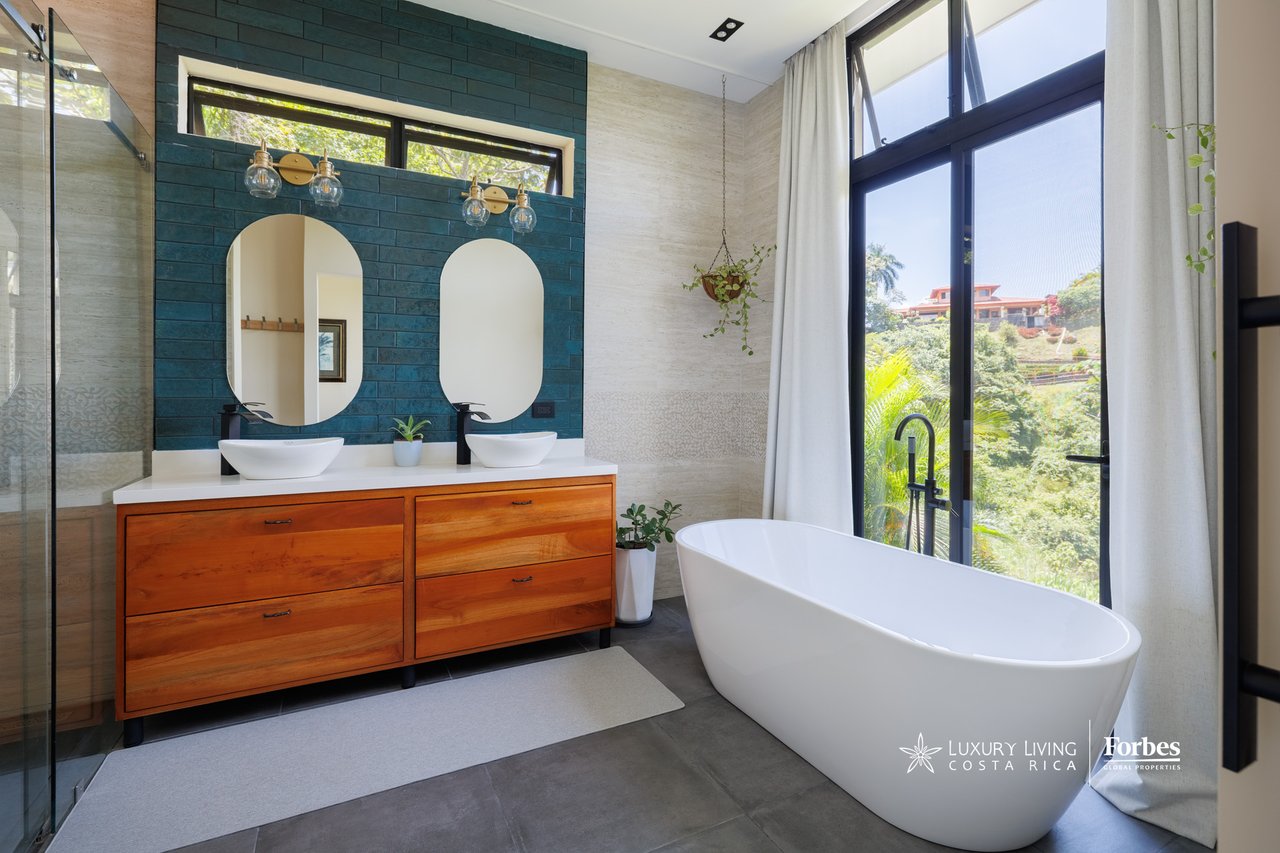 Modern bathroom with a freestanding tub, double wooden vanity, and large windows overlooking greenery.