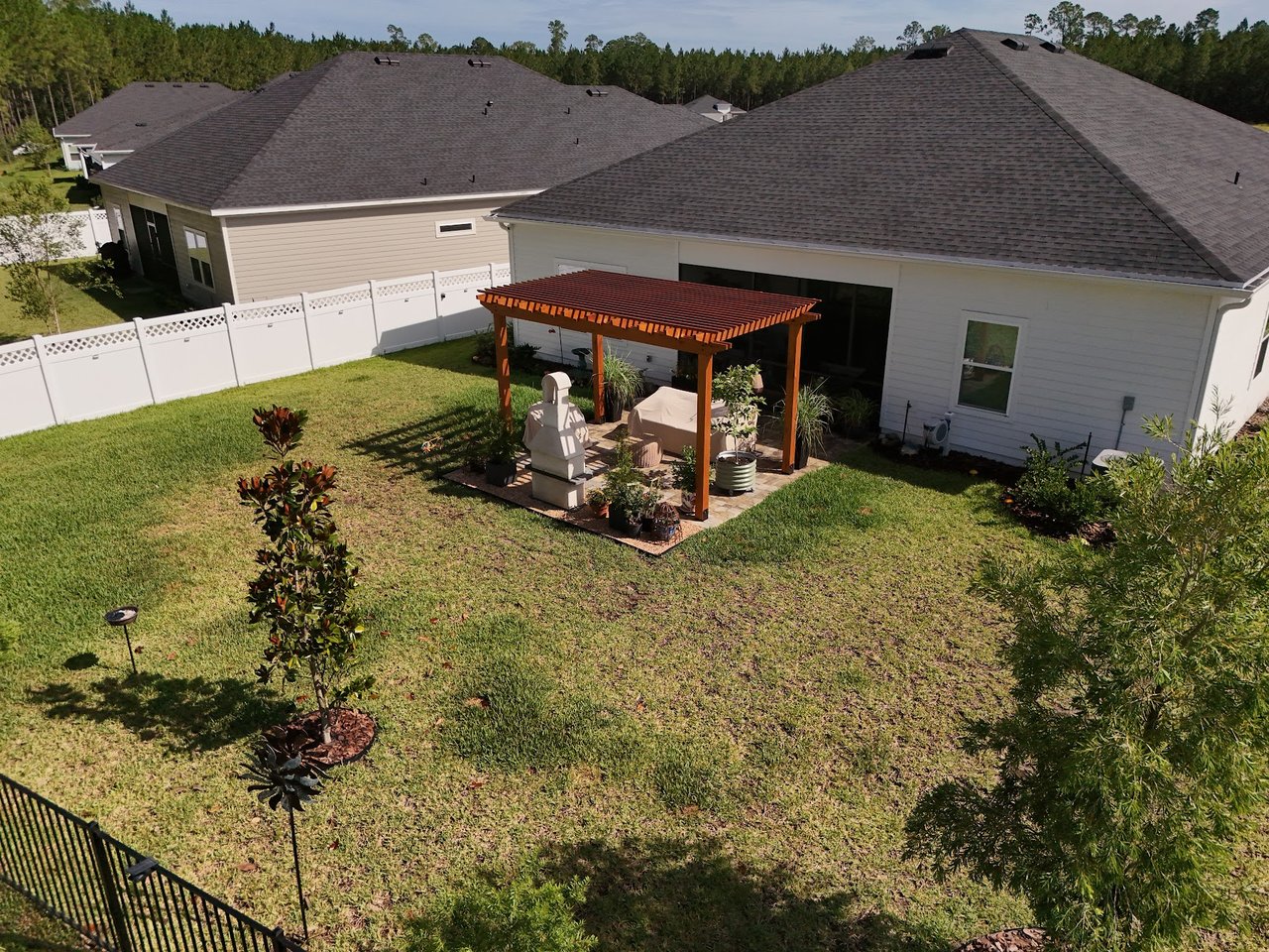 Aerial view of a home’s backyard with a wooden pergola, patio seating, and fenced lawn.