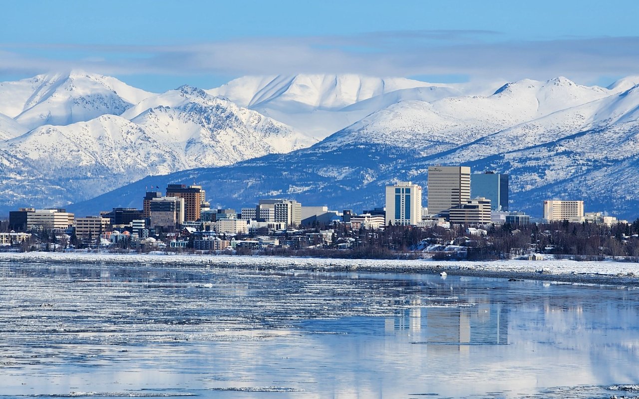 Anchorage skyline beneath snow-covered mountains, reflected in icy water.