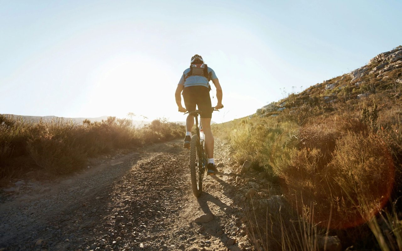 A cyclist pedals uphill on a rocky trail through sunlit desert scrub.