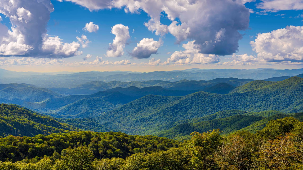 Panoramic forested mountain ridges beneath a blue sky filled with large clouds.