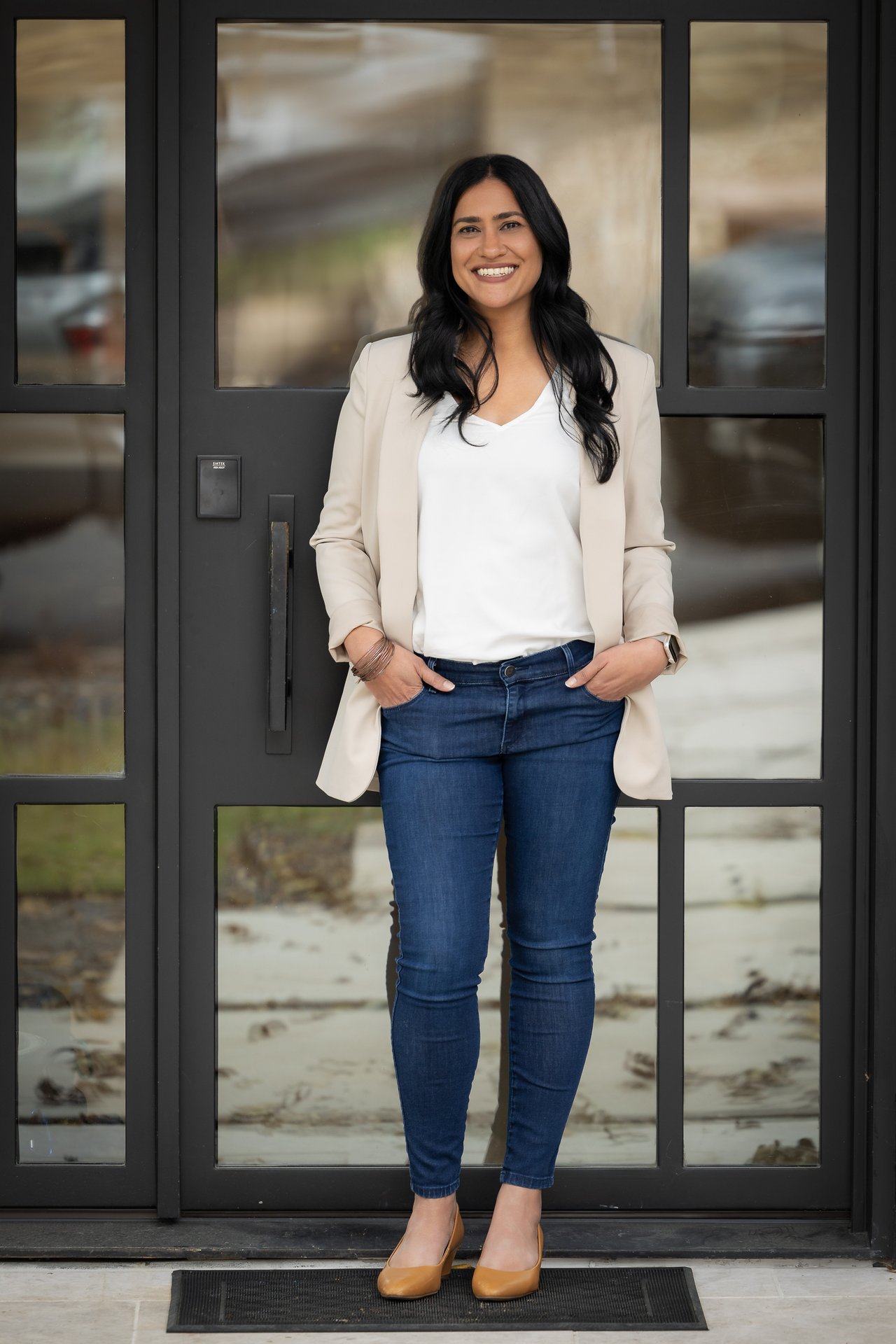 Sonia Allison stands smiling in a light blazer and jeans before a glass-paneled entrance.