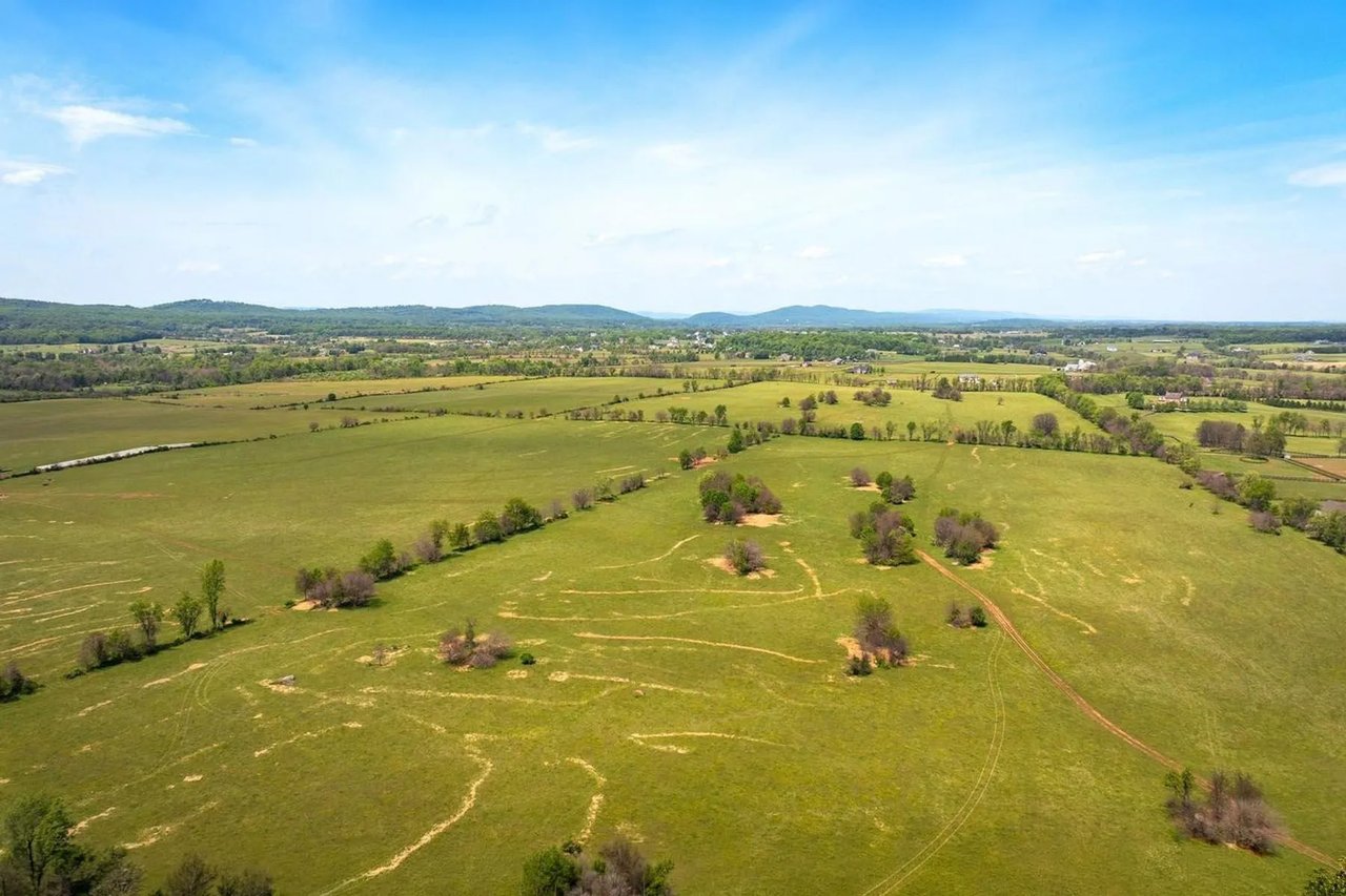 Aerial pastureland with scattered trees and dirt tracks, bordered by farmland and distant wooded hills.