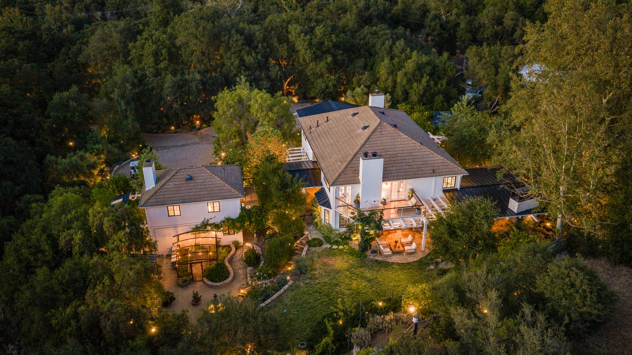 Aerial view of two wooded homes with landscaped grounds and an illuminated patio at dusk.