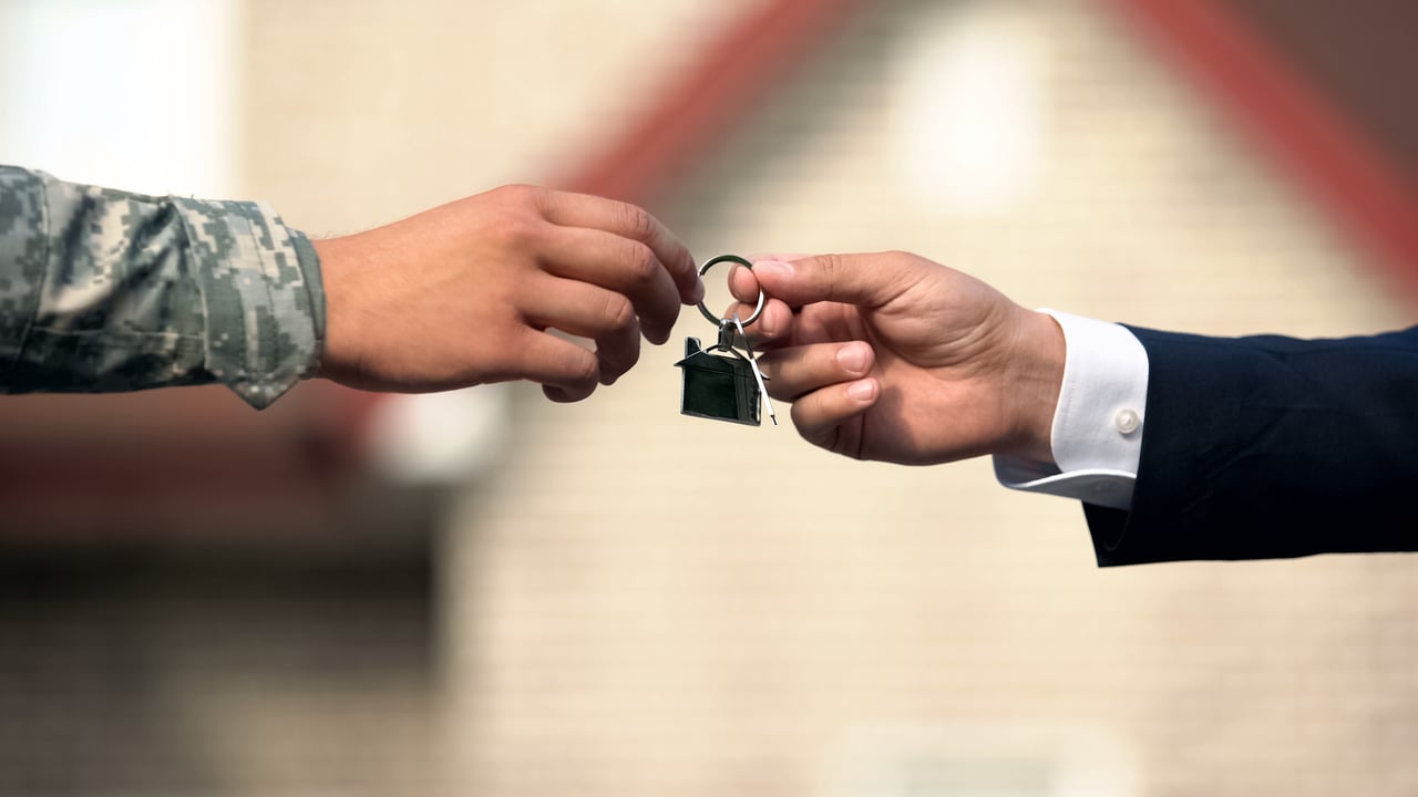 Hands exchange house keys, one in a camouflage sleeve and the other in a dark business suit.