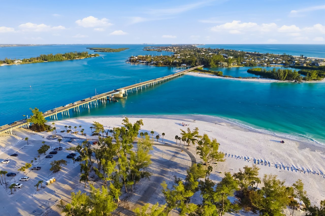 Aerial view of a bridge over turquoise water beside a white-sand beach and coastal homes.