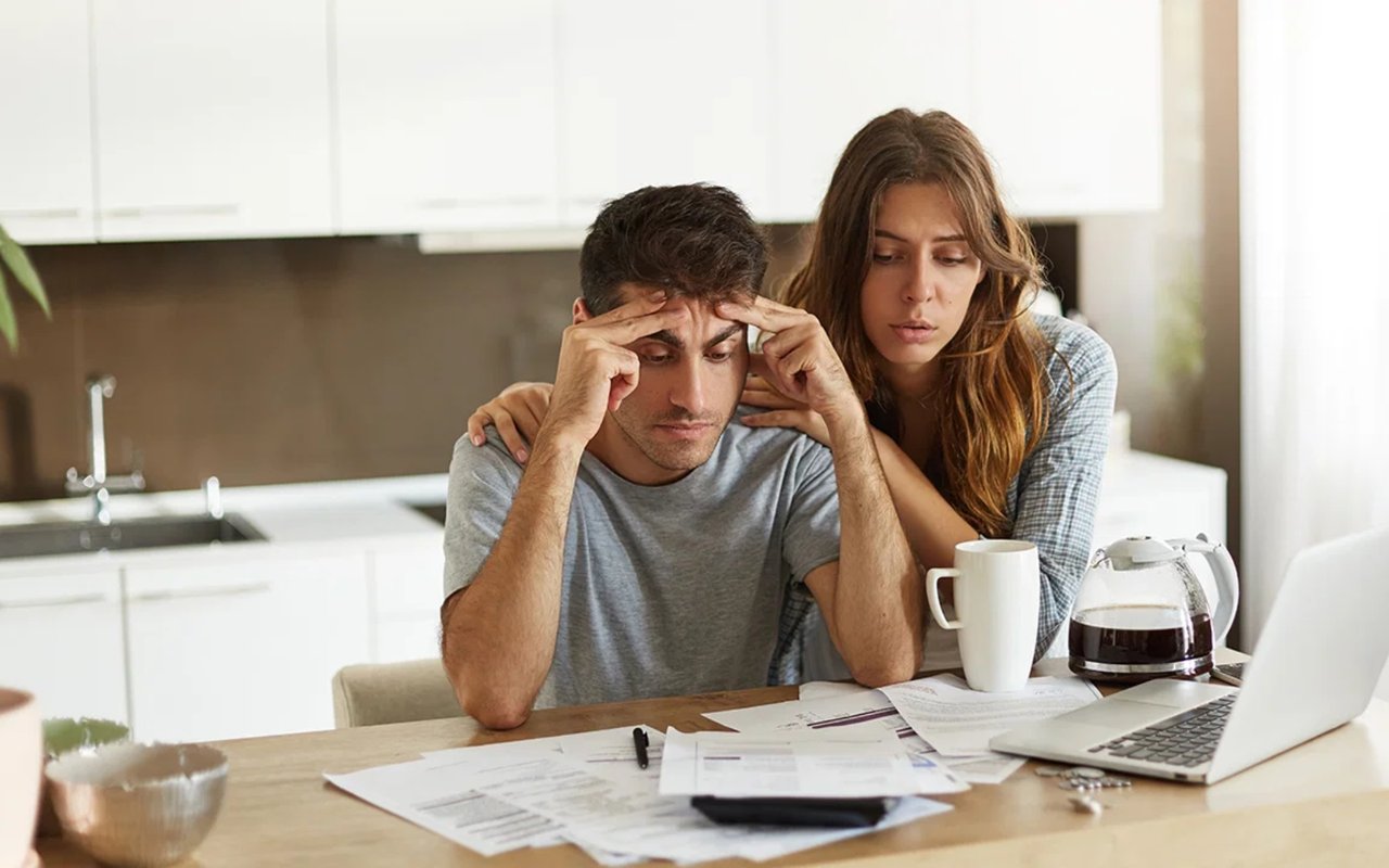 A worried man reviews bills at a kitchen counter as a woman offers support.