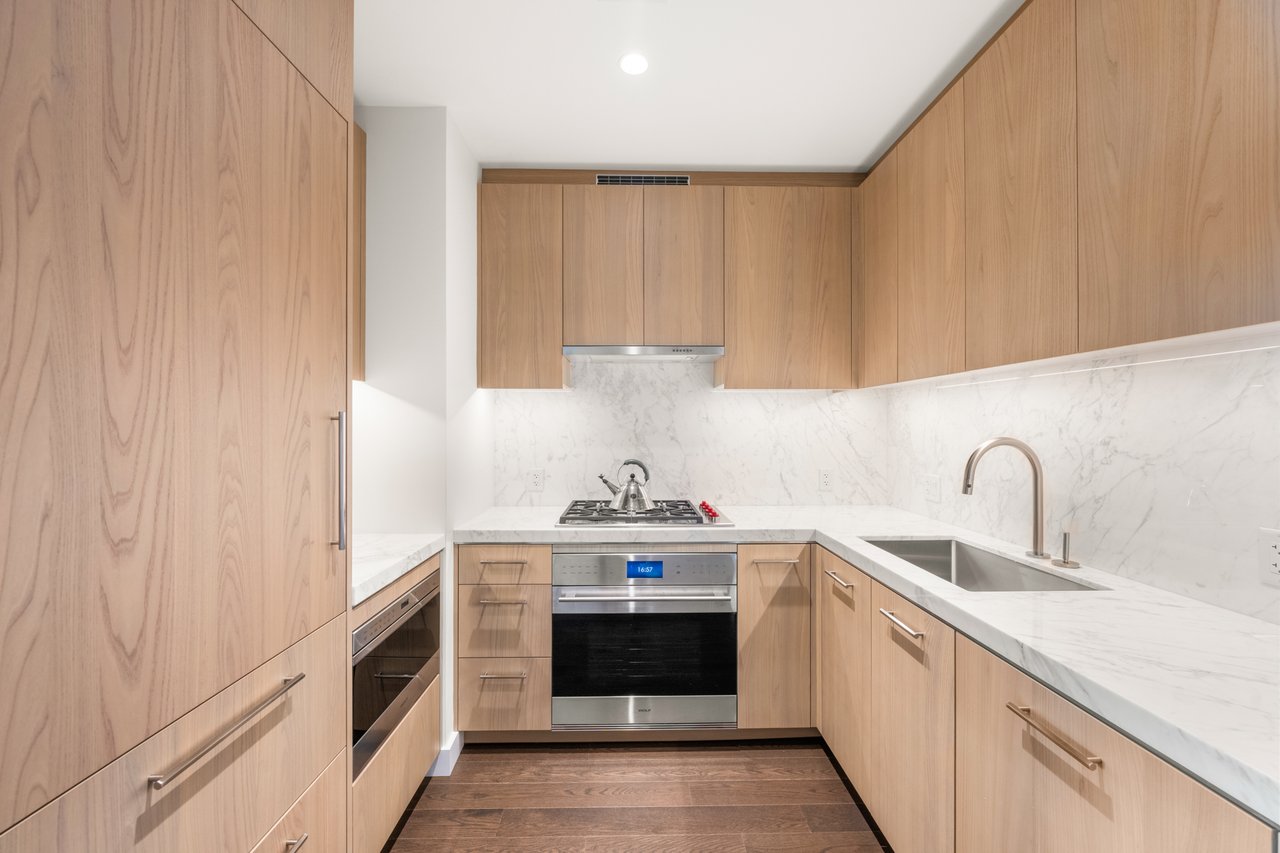 Modern kitchen with light wood cabinets, marble counters, and stainless steel appliances.