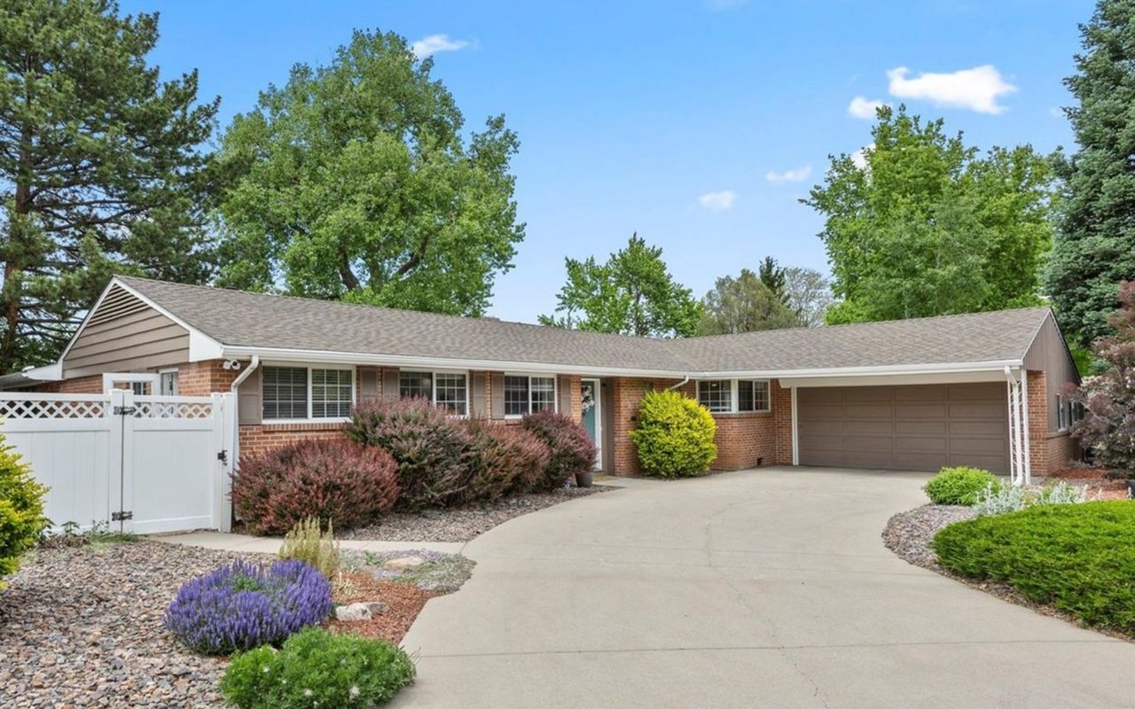 Front exterior of a sold single-story brick ranch home in Denver with a two-car garage, curved concrete driveway, xeriscaped front yard with purple sage, and mature trees under a blue sky.