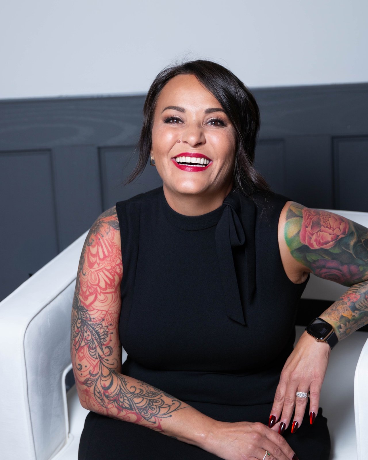 Nicole Hagedorn Smith seated in a white chair, smiling, with tattooed arms.