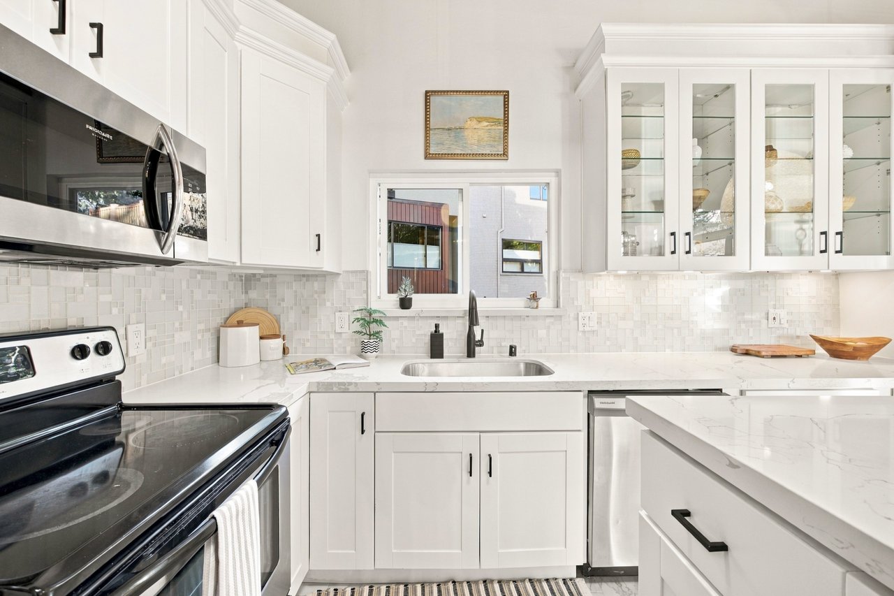 Bright white kitchen with marble counters, glass-front cabinets, and a window above the sink.