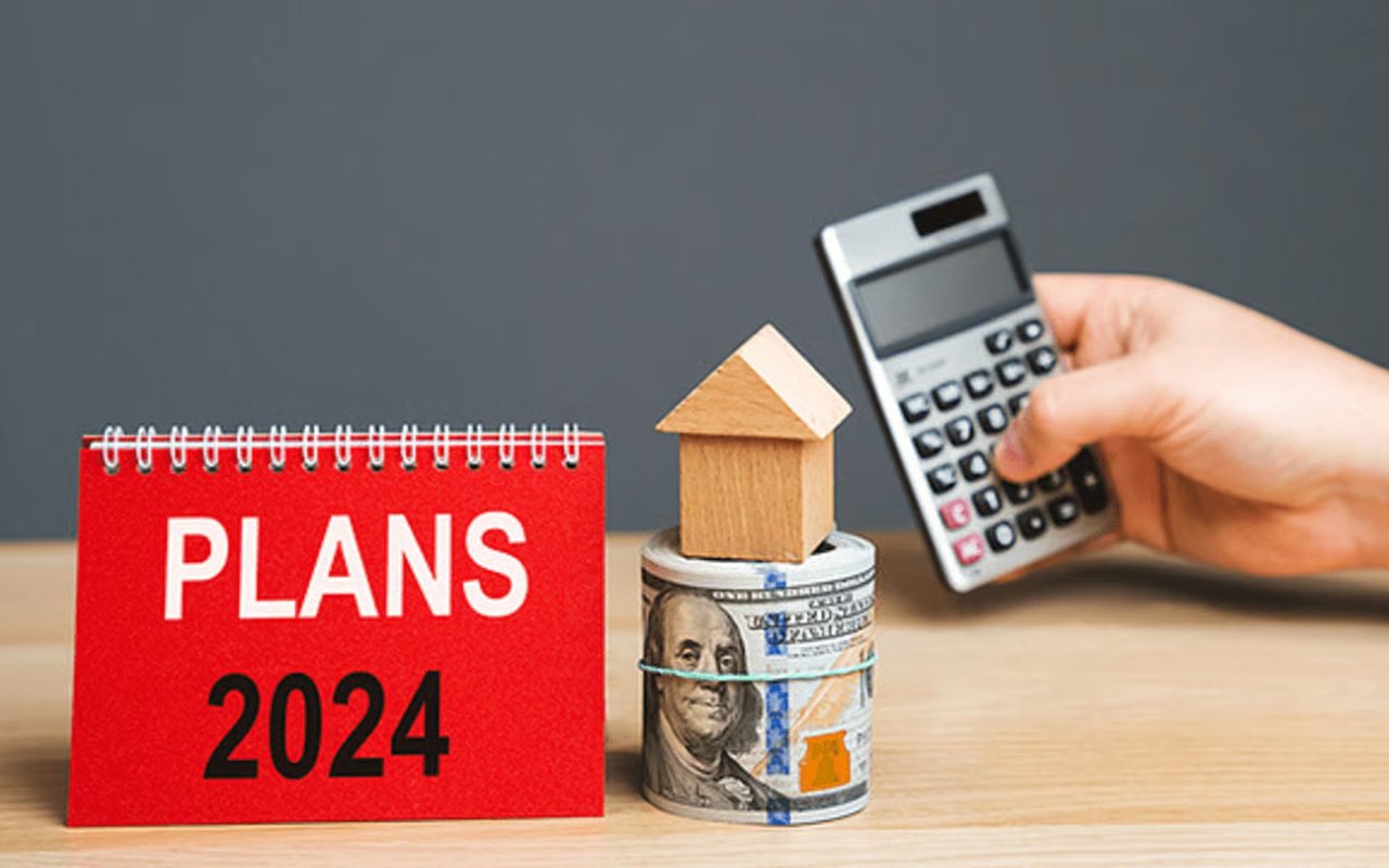 A wooden model house sits on rolled cash beside a 2024 planning calendar and calculator.