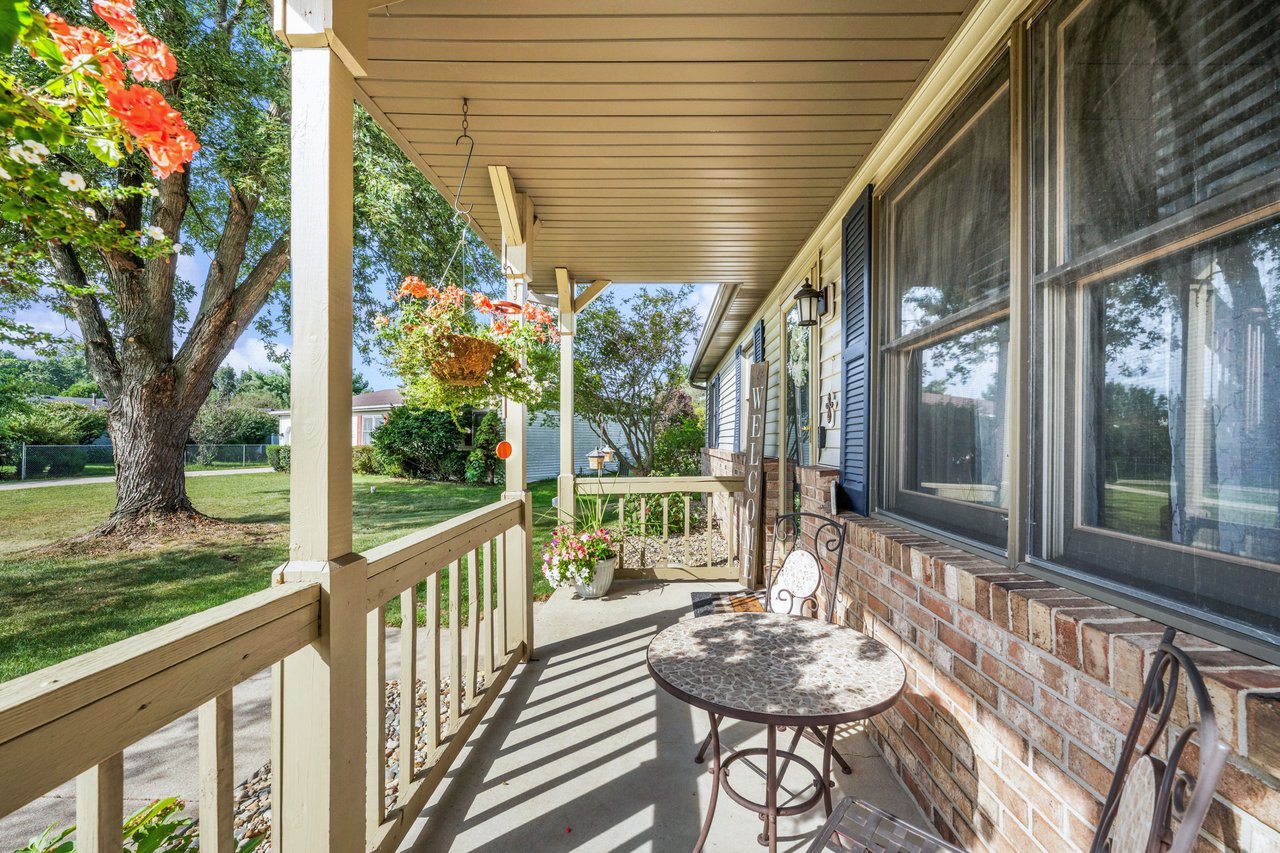 Covered front porch with brick wall, wooden railings, hanging flowers, and a round patio table.