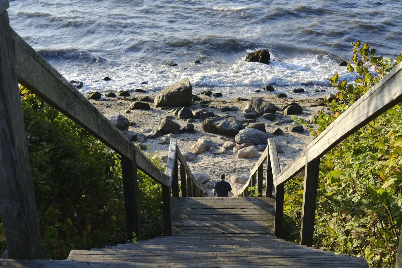 Wooden stairs lead down to a rocky beach and choppy surf, with a person descending.