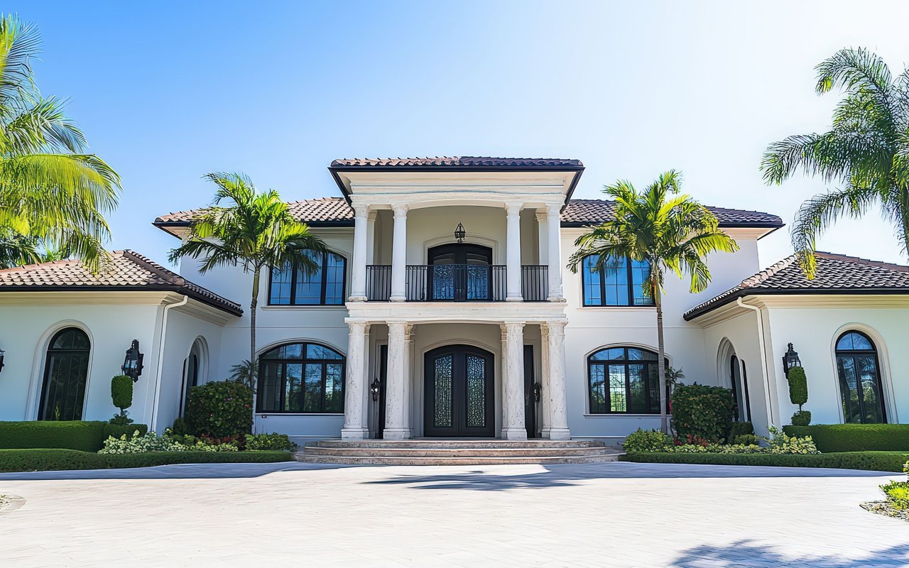 Large white two-story house with a columned entrance, tiled roof, and palm trees.