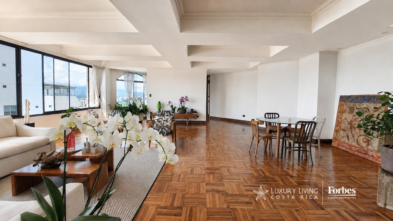 Furnished condominium living and dining room with parquet floors, orchids, and city views through large windows.