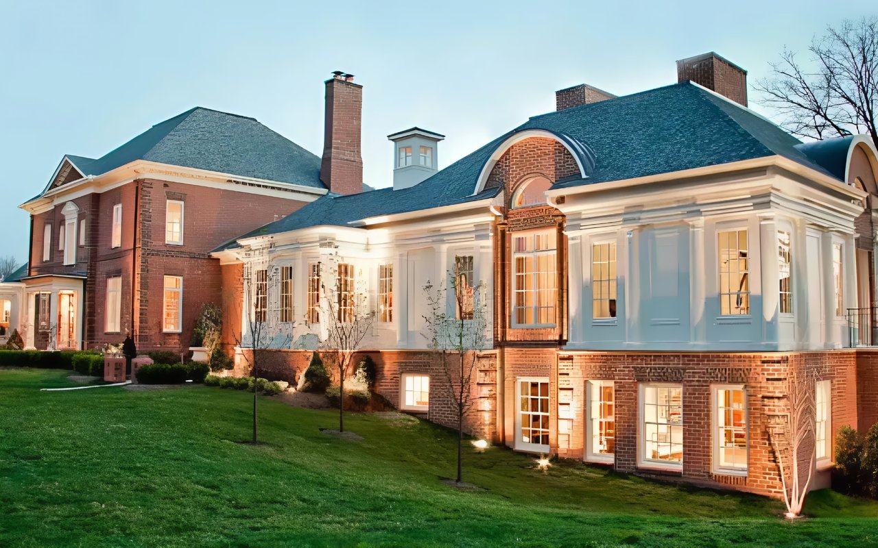 Large brick house with a white facade, arched entry, and warmly lit windows at dusk.