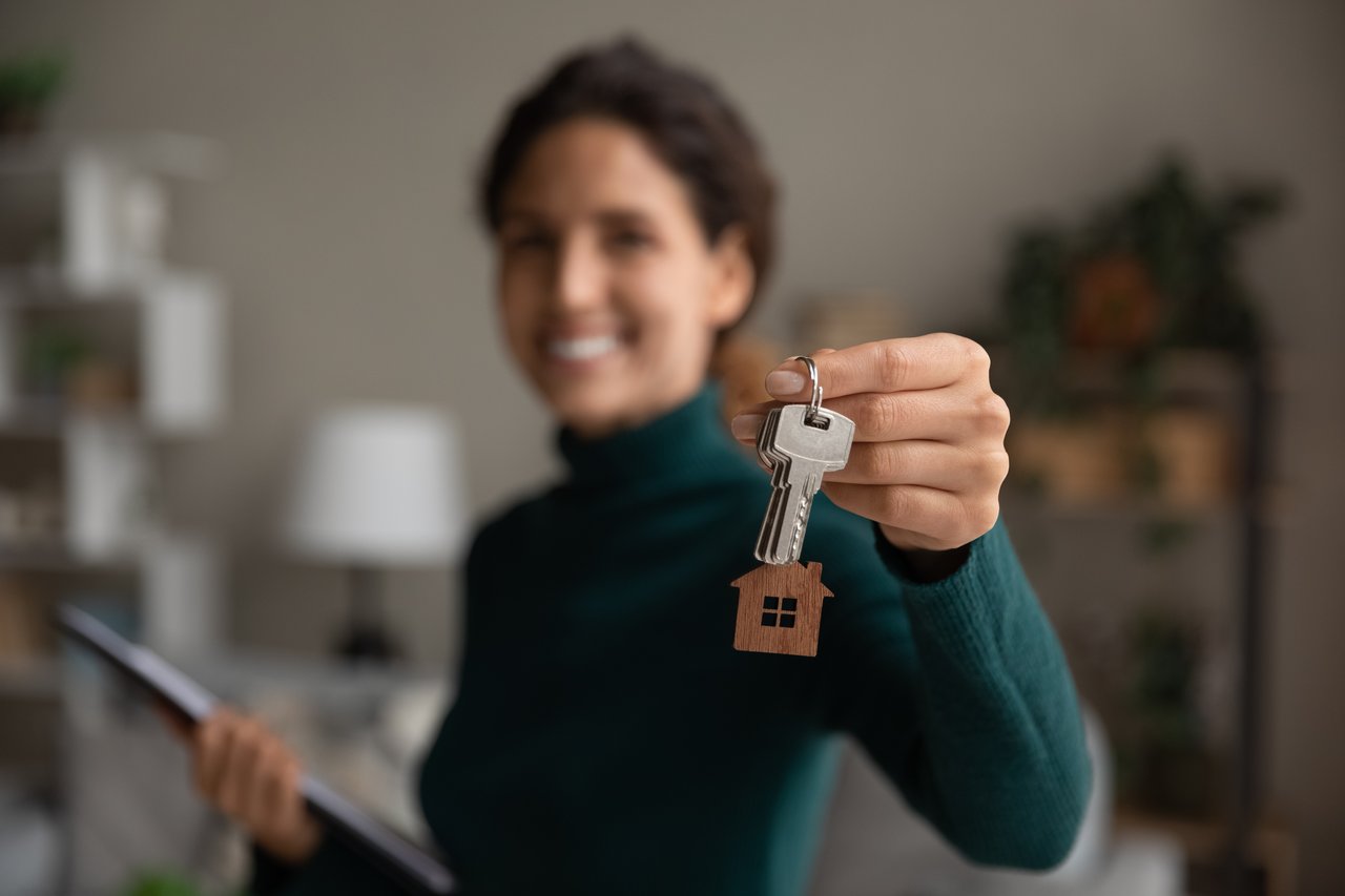 Smiling woman holding a house key with a home-shaped keychain in a cozy living room.