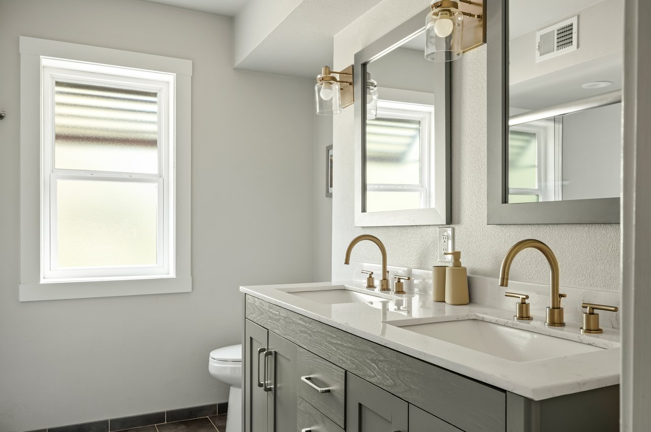 Bathroom double vanity with brass faucets, gray cabinetry, large mirrors, and a window.