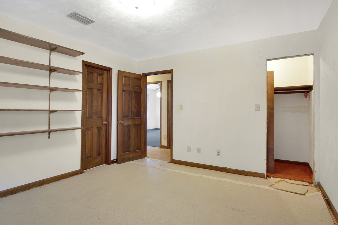 Empty room with wooden shelves on the left wall, two wooden doors, and an open closet with red carpet. The walls and floor are bare, suggesting renovation.