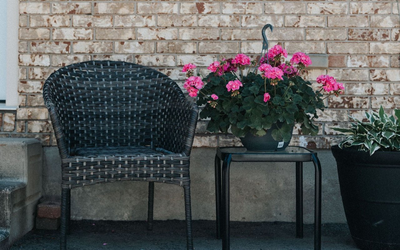 Patio with a dark wicker chair, pink flowers on a table, and a brick wall.