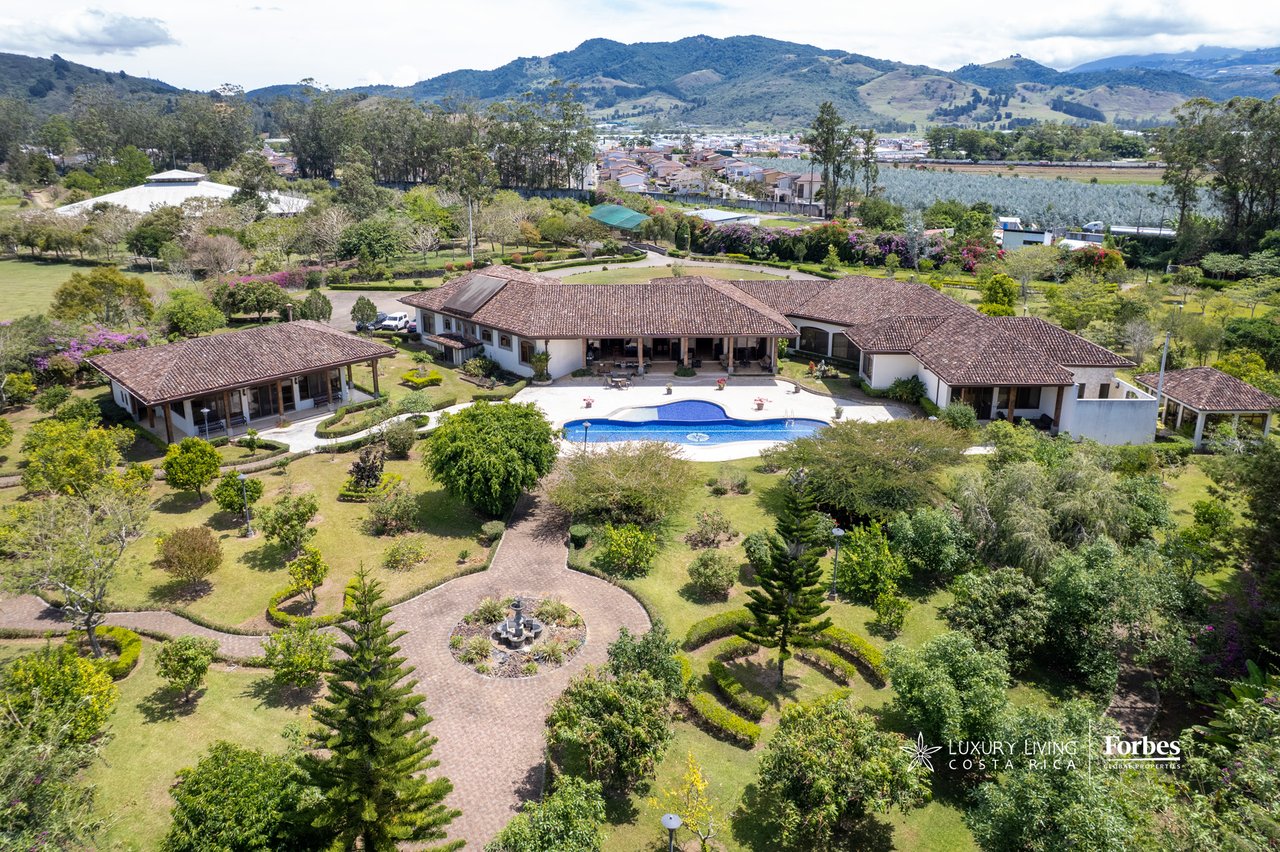 Aerial view of a tiled-roof estate with a central pool, landscaped grounds, and mountains beyond.