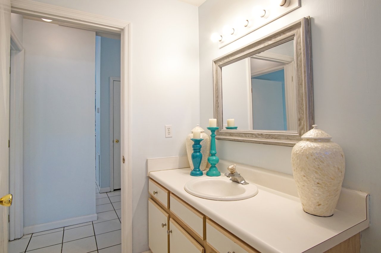 Bright bathroom with a white countertop and sink, large mirror, teal candle holders, and decorative vase. Soft lighting creates a calm atmosphere.