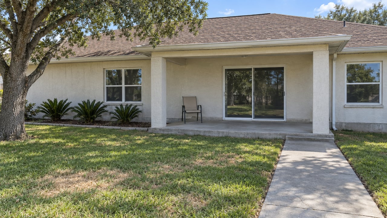 Single-story stucco home with a covered rear concrete patio, sun-faded brown shingle roof, patchy lawn and mature spreading oak.