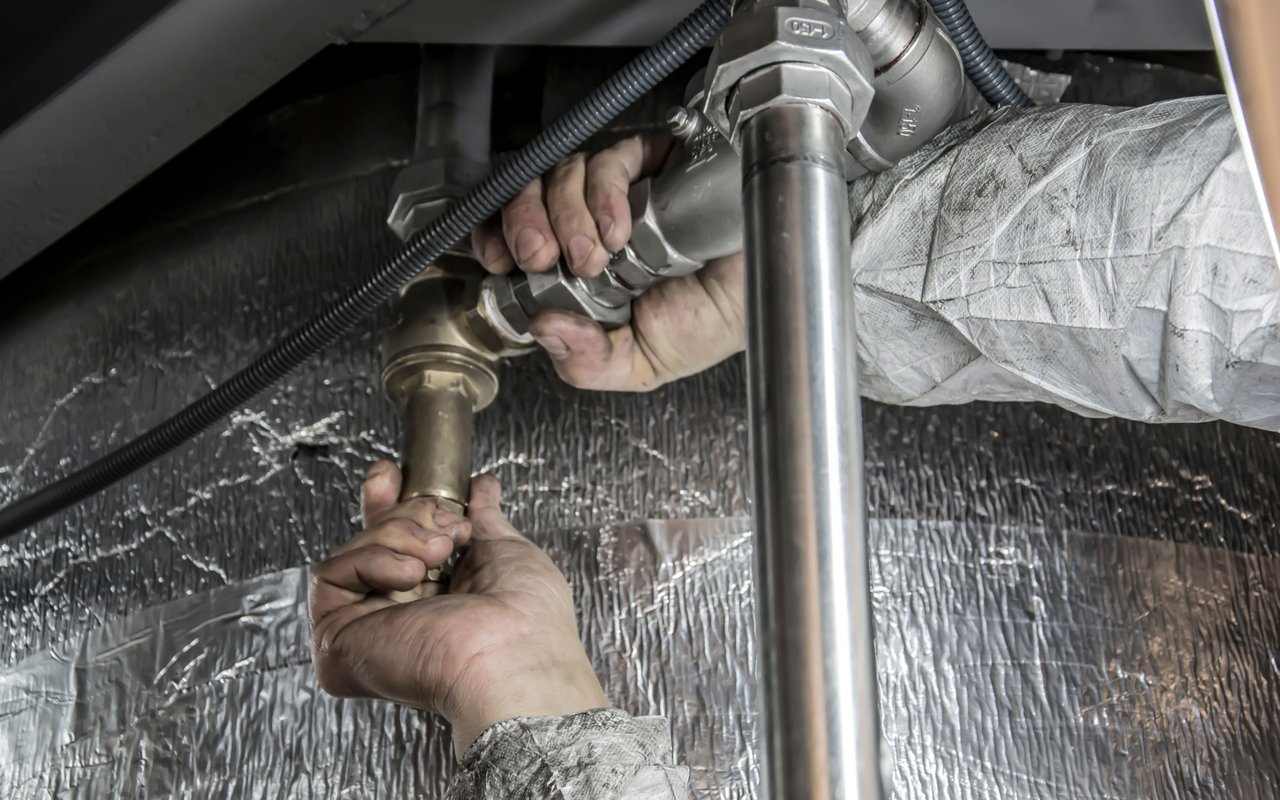Worker connecting plumbing pipes beneath a home, surrounded by reflective insulation and wiring.