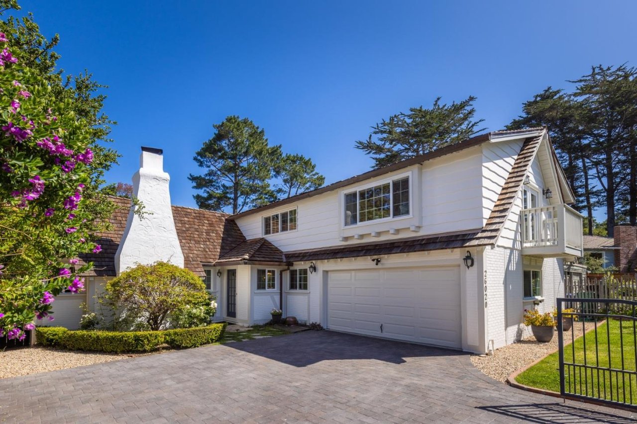 White two-story house with a double garage, upper balcony, and flowering shrubs beside the driveway.