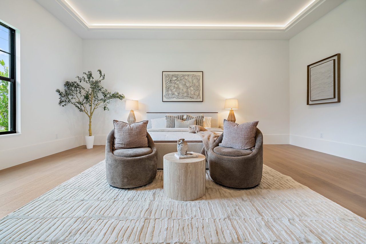 Staged bedroom with a tray ceiling, upholstered chairs, and a large textured rug.