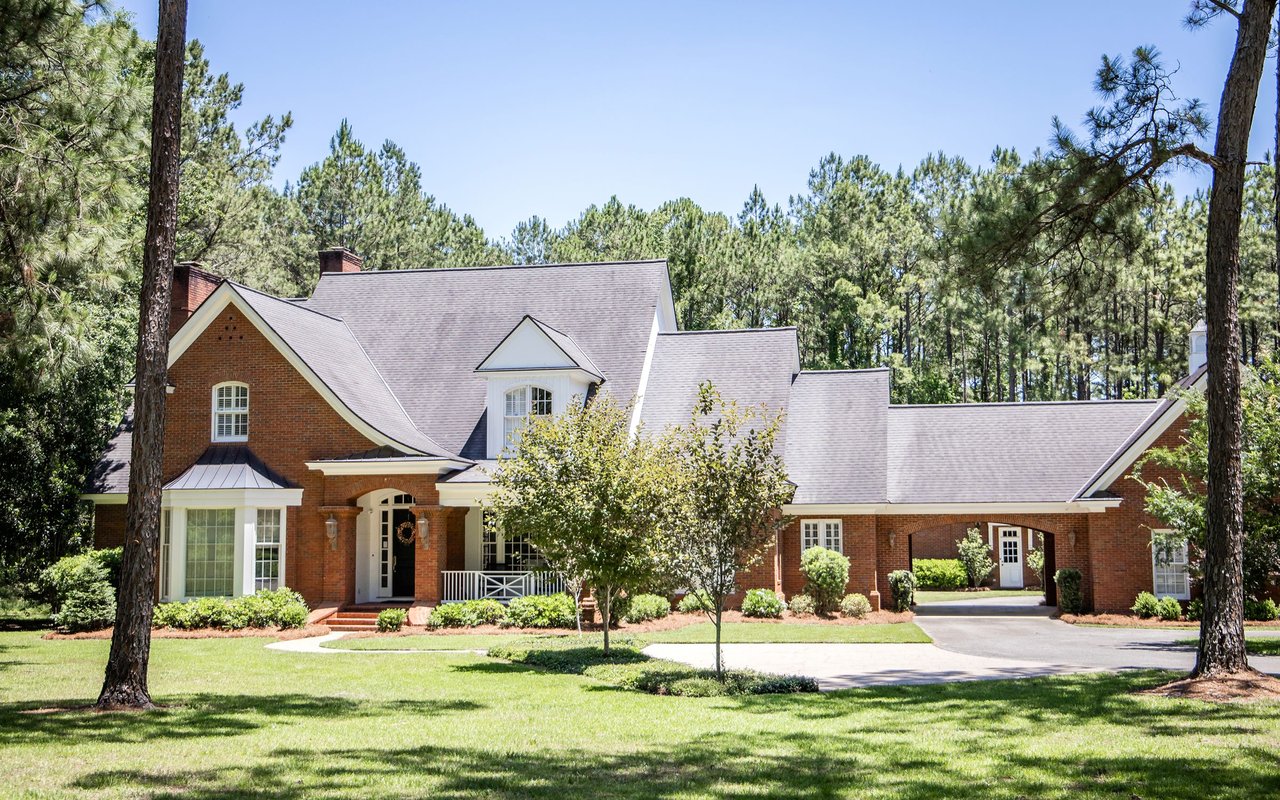 Large brick house with multiple gabled roofs, an attached garage, and a tree-lined lawn.