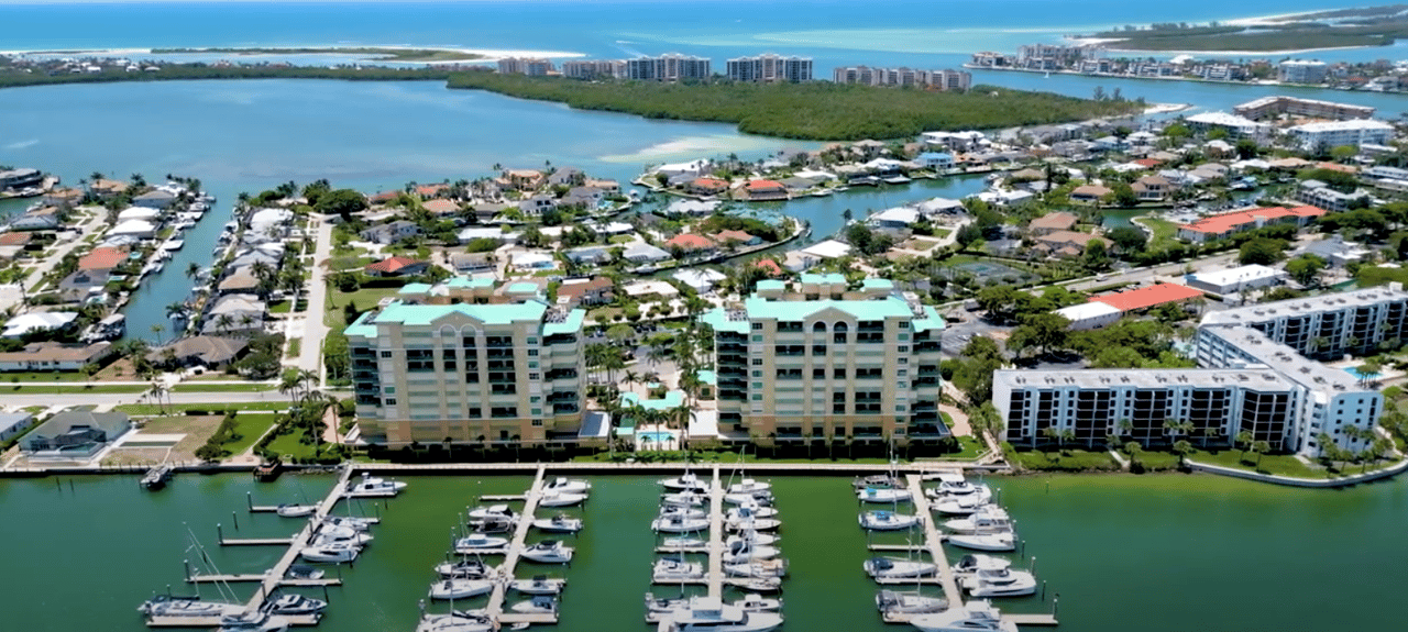 Aerial marina with rows of boats in front of waterfront condominiums and homes.