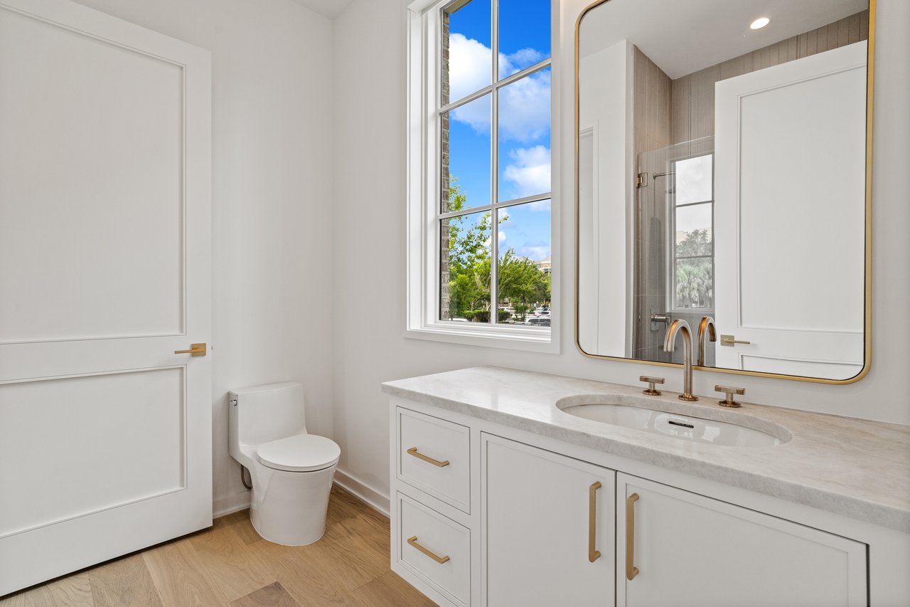 Bright bathroom with a white vanity, brass fixtures, large mirror, and tall window.