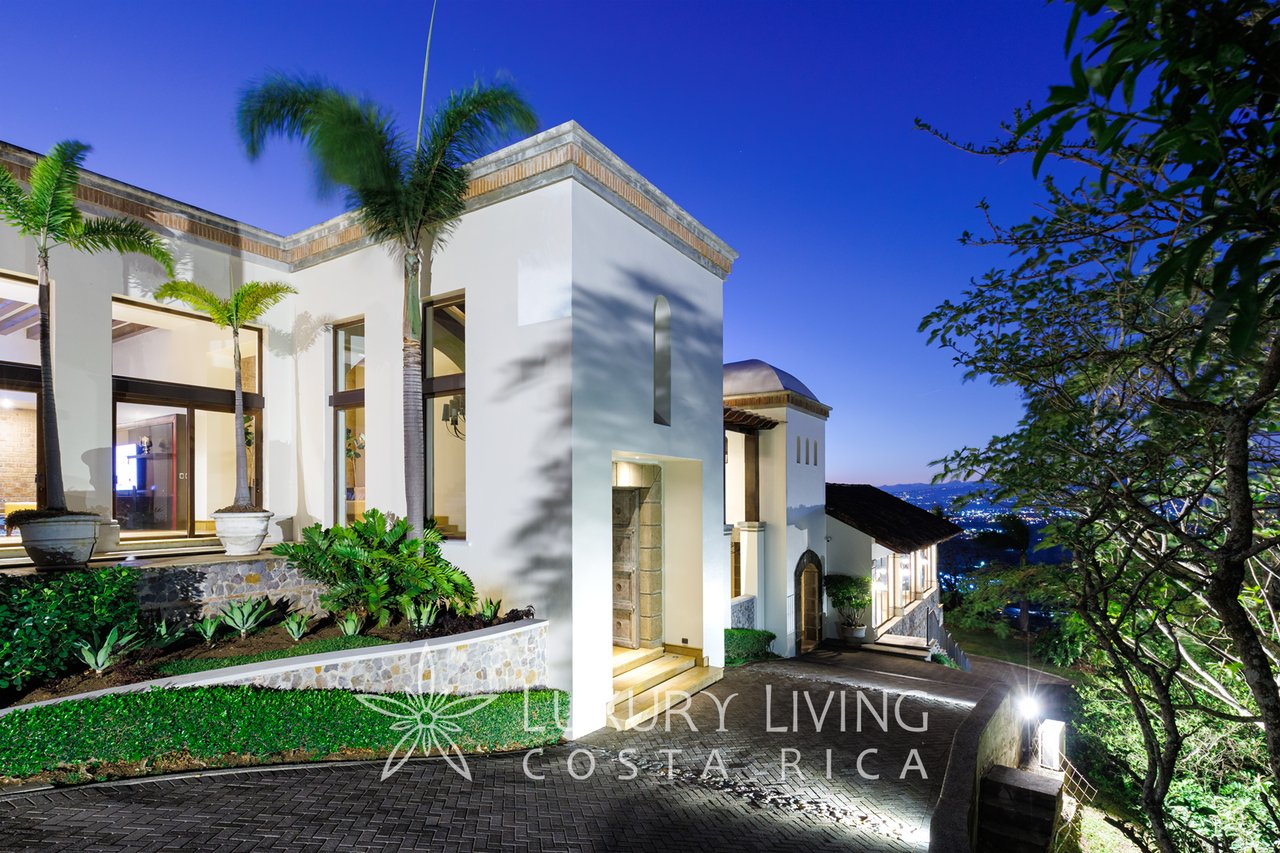 White luxury home with palm trees and landscaped grounds, illuminated against a hillside with distant city lights.