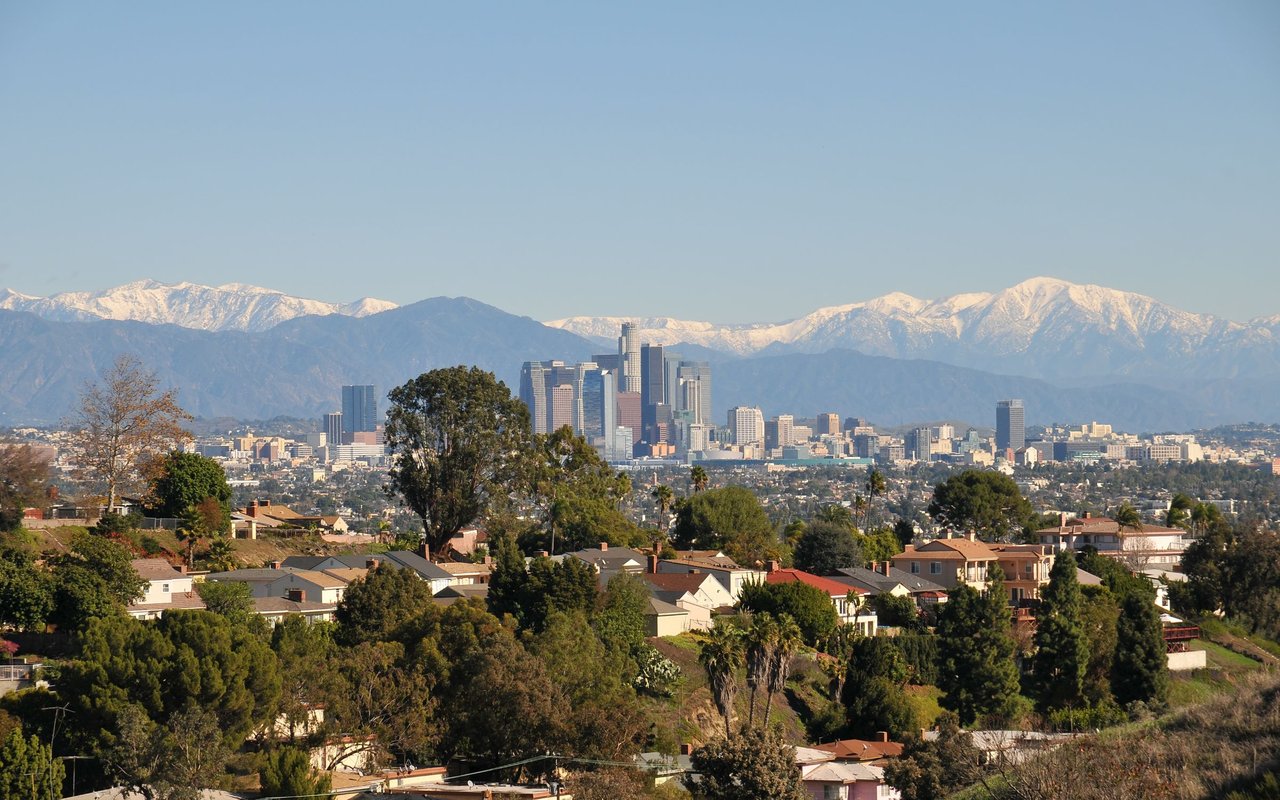 Downtown Los Angeles skyline beyond hillside homes, with snow-covered mountains in the distance.