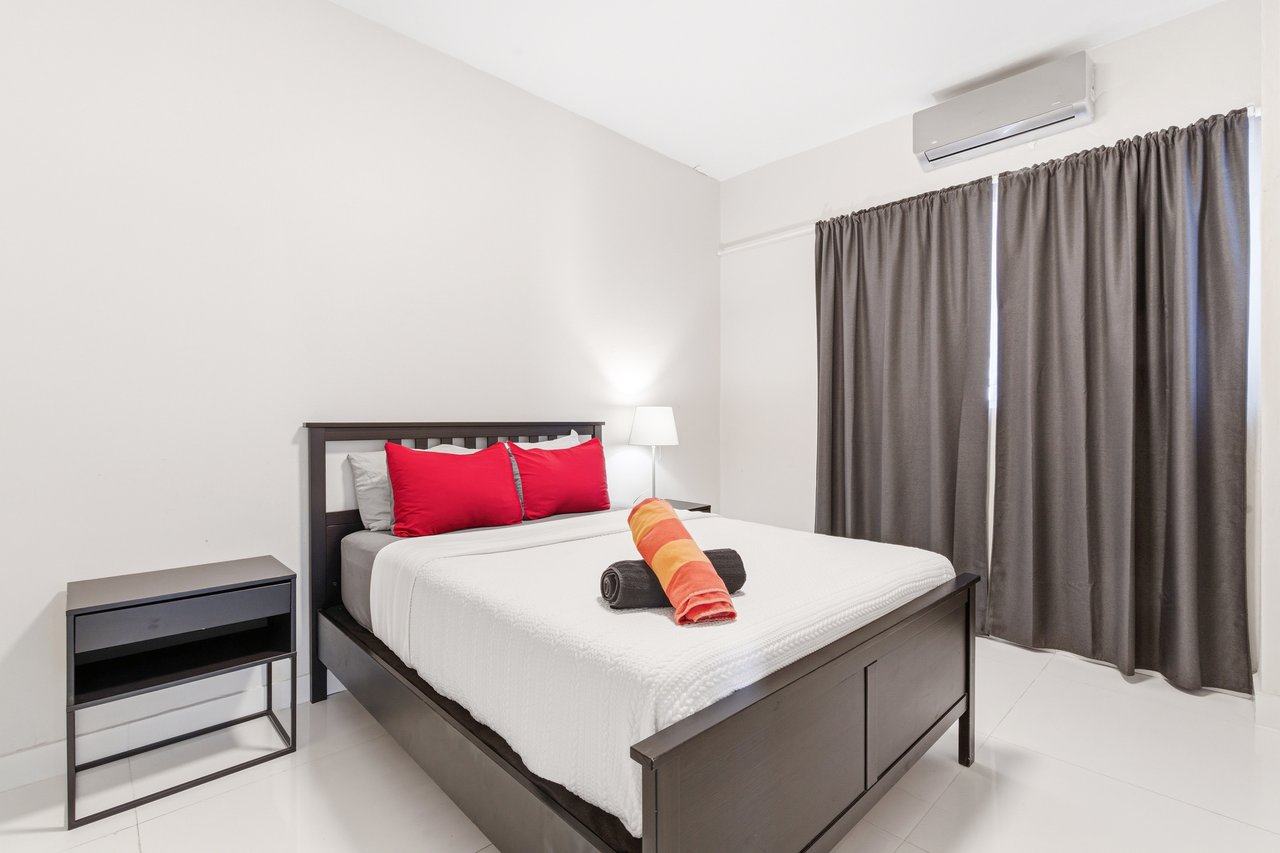 Furnished bedroom with red pillows, folded towels on the bed, and gray curtains.