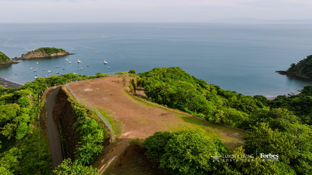 Cleared residential lot above a sheltered bay, surrounded by tropical greenery with boats offshore.