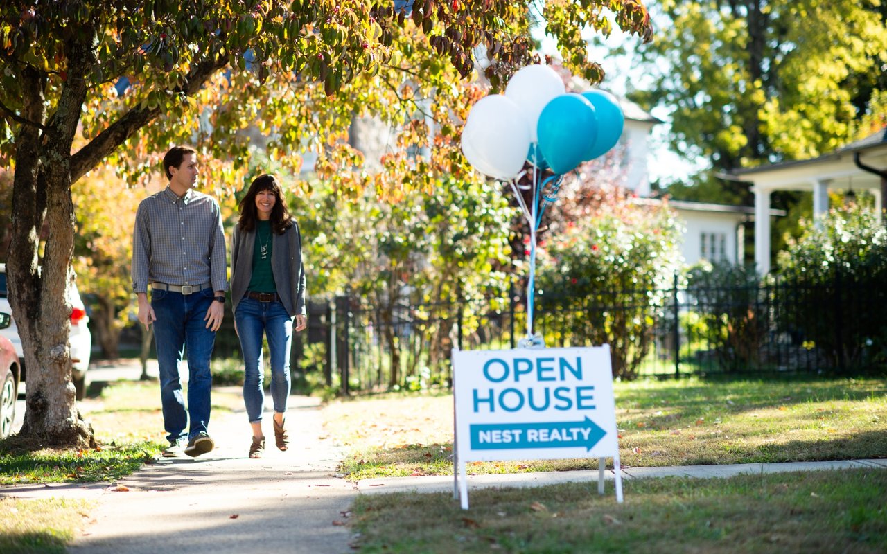 A couple walks toward a blue-and-white open house sign beneath autumn trees.