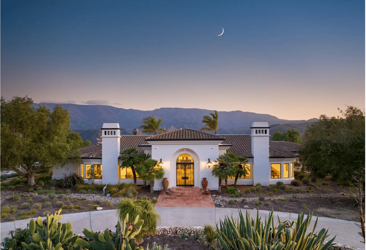 White Mediterranean-style home with an arched entry, desert landscaping, and mountain views at dusk.
