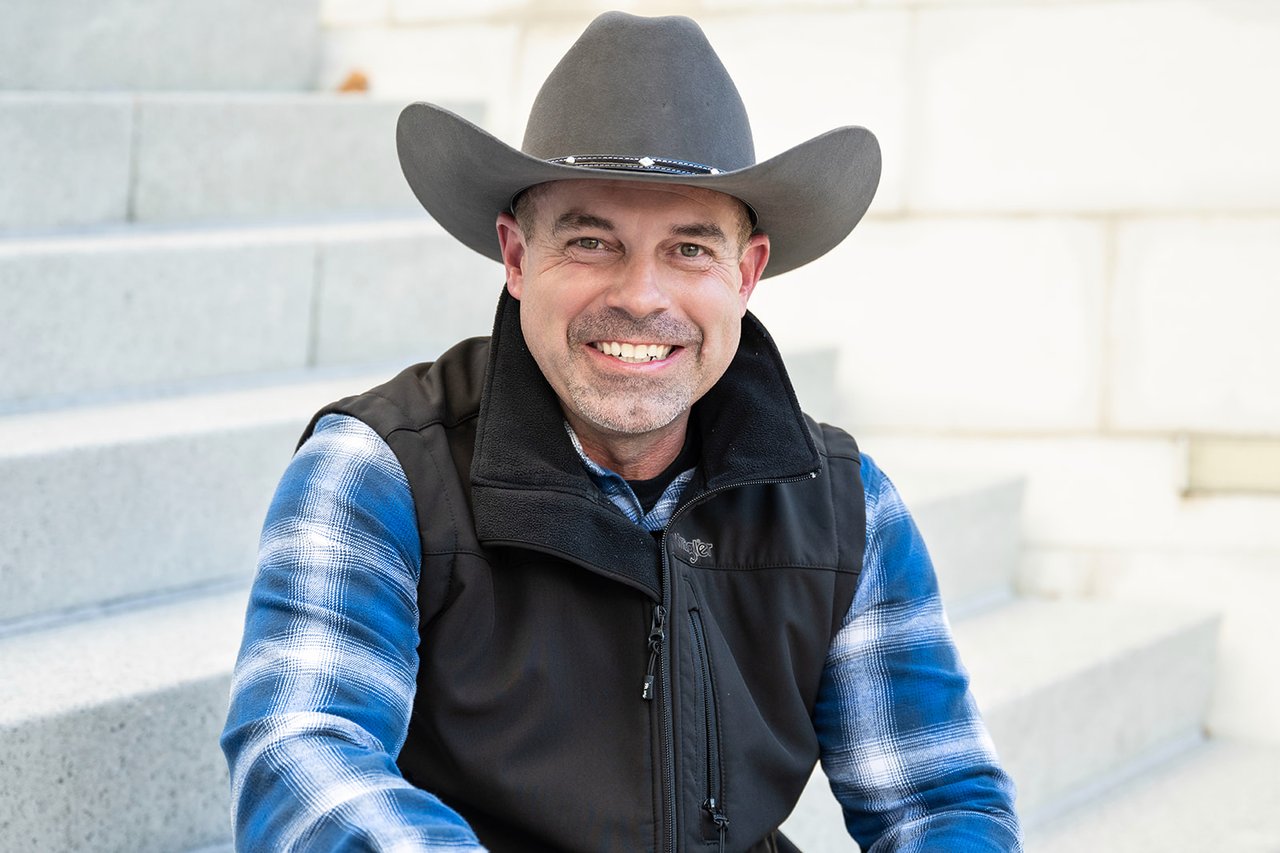 Scott Stamper smiling in a cowboy hat, blue plaid shirt, and black vest.