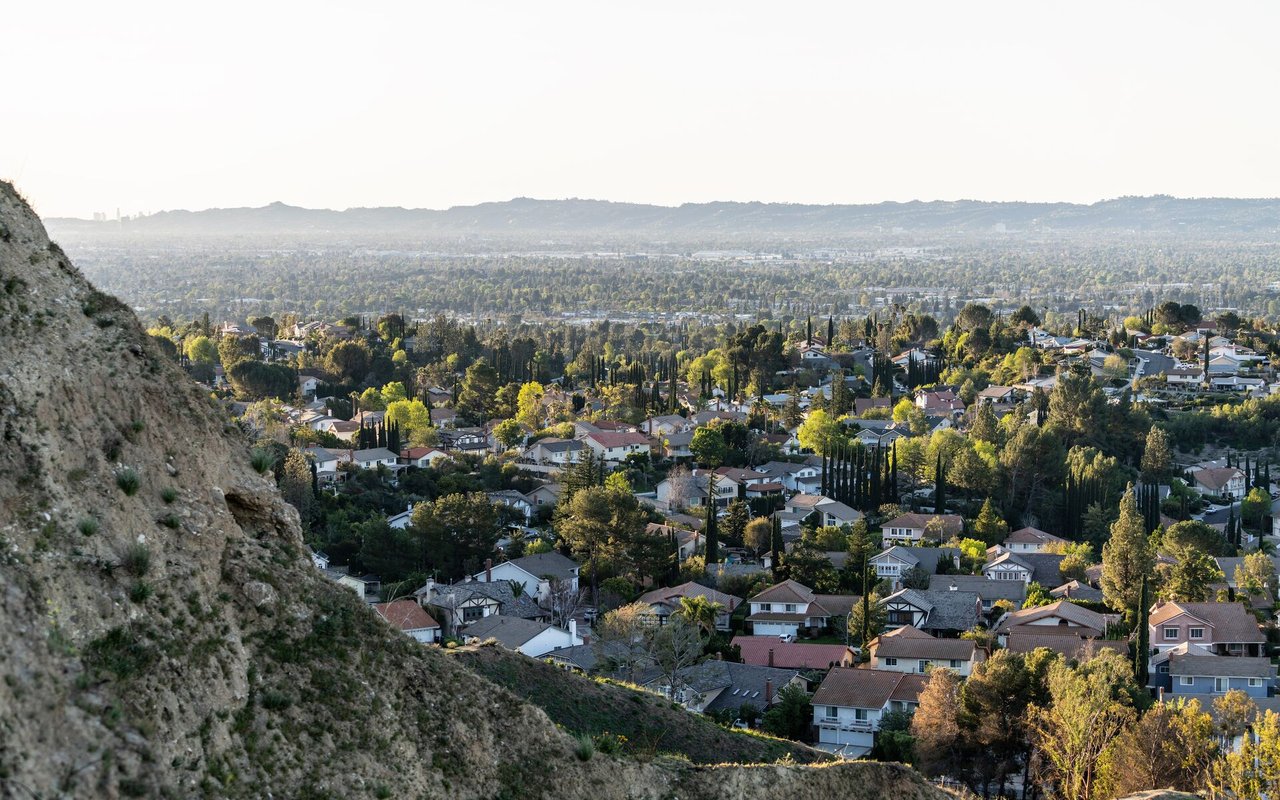 Residential neighborhood spreads across a green valley below distant hills, viewed from a rocky hillside.