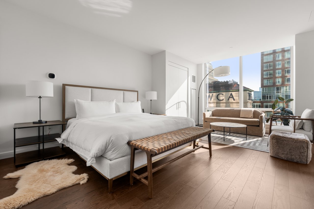 Furnished bedroom with a large white bed, woven bench, and sitting area beside floor-to-ceiling windows.