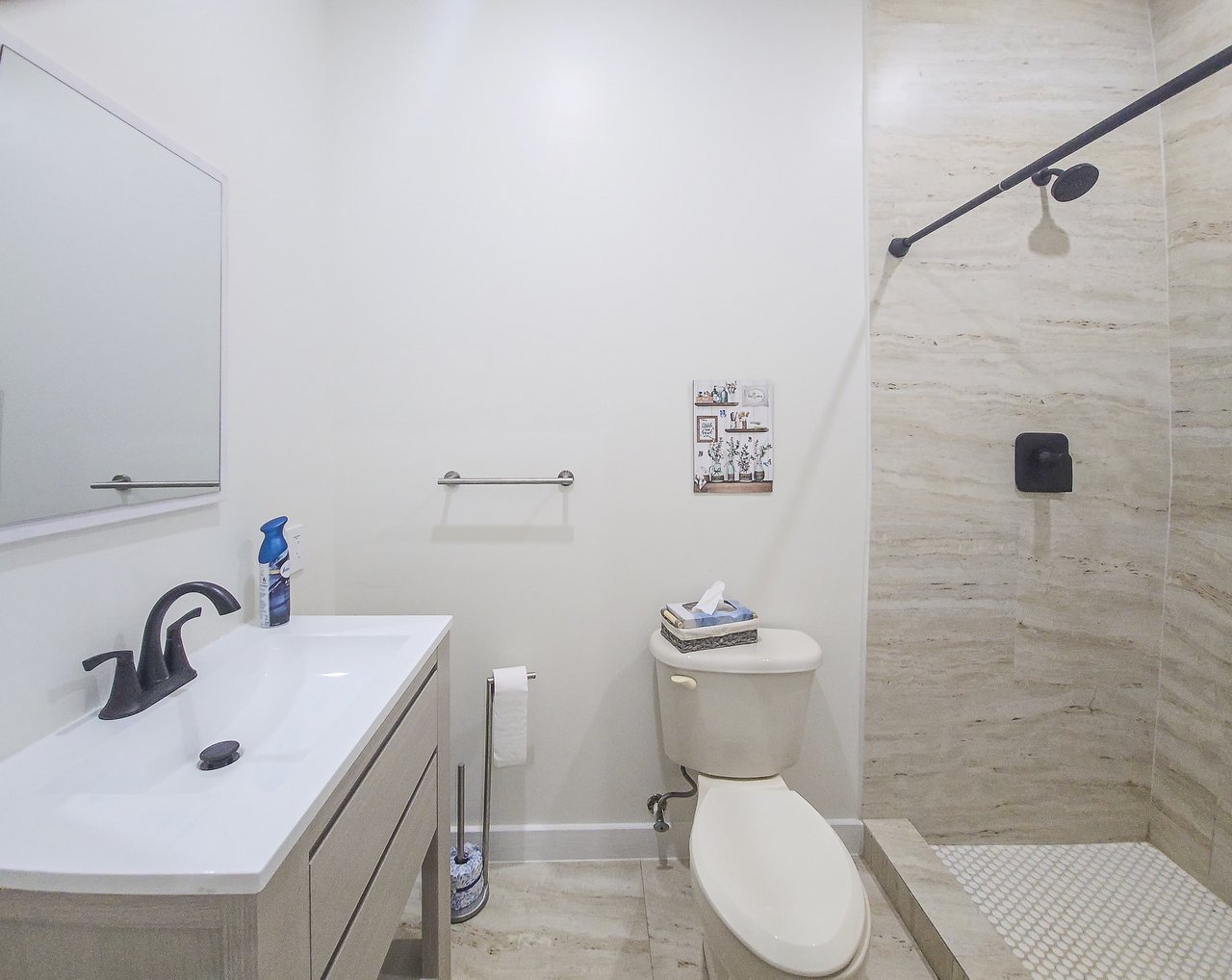 Bathroom with a white vanity, black faucet, toilet, and beige stone-tiled walk-in shower.