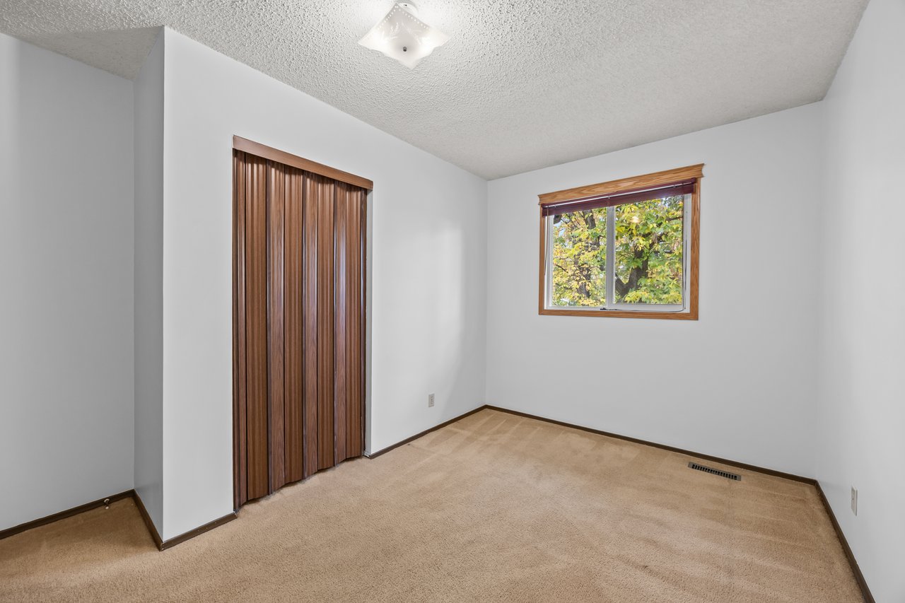 Unfurnished bedroom with beige carpet, brown sliding closet doors, and a window overlooking leafy trees.