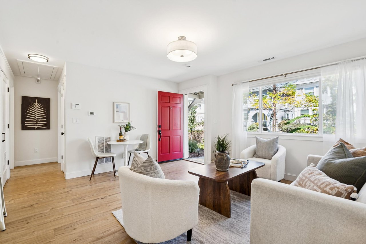 Bright staged living room with a striking red front door, white seating, and hardwood floors.